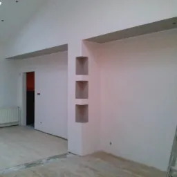 Wykończone wnętrze: białe ściany z wnękami, jasna podłoga, grzejnik, widoczne fragmenty drabiny i okna dachowego. Minimalistyczny design z elementami dekoracyjnymi.