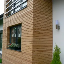 Elewacja domu z drewnianą okładziną, widok z zewnątrz. Okno z kwiatami, nowoczesna lampa ścienna i fragment ogrodu.