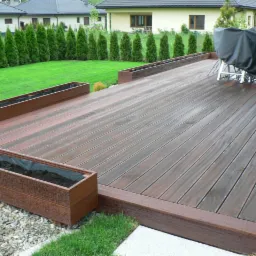 Nowoczesny, brązowy taras z desek kompozytowych. Widoczne donice z ziemią, trawnik i rząd tuj w tle. Zadaszony grill na tarasie.