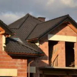 Surowy budynek w trakcie budowy z widocznym dachem pokrytym ciemną dachówką, fragmenty drewnianej konstrukcji dachowej i ceglane ściany bez okien.