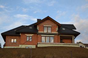 Dom jednorodzinny w trakcie budowy, z widocznym nowym, czarnym pokryciem na dachu i oknami dachowymi, częściowo otynkowany, z elementami z czerwonej cegły, usytuowany na wzniesieniu.