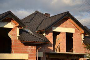 Surowy budynek w trakcie budowy z widocznym dachem pokrytym ciemną dachówką, fragmenty drewnianej konstrukcji dachowej i ceglane ściany bez okien.