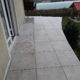Balkon z jasnymi, kwadratowymi płytkami ceramicznymi, widoczne ślady po butach, w tle ogród z altaną.