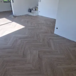 Ułożona parkietowa podłoga w jodełkę w jasnym pokoju z białymi ścianami i schodami. Widoczne narzędzia i materiały malarskie. Surowy betonowy strop.