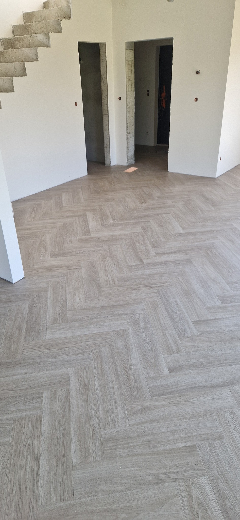 Ułożona podłoga z paneli w jodełkę, jasne odcienie drewna, surowe ściany i schody w tle, wnętrze w trakcie wykańczania.