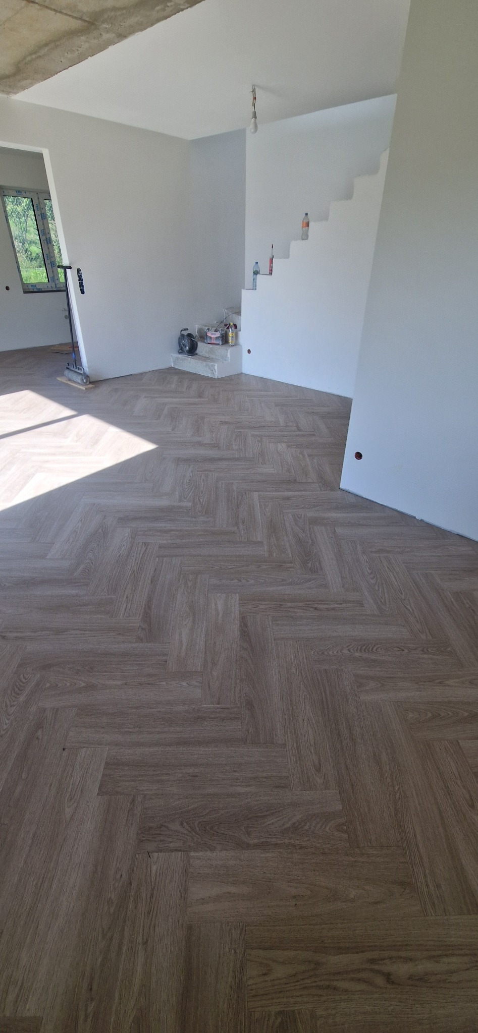 Ułożona parkietowa podłoga w jodełkę w jasnym pokoju z białymi ścianami i schodami. Widoczne narzędzia i materiały malarskie. Surowy betonowy strop.