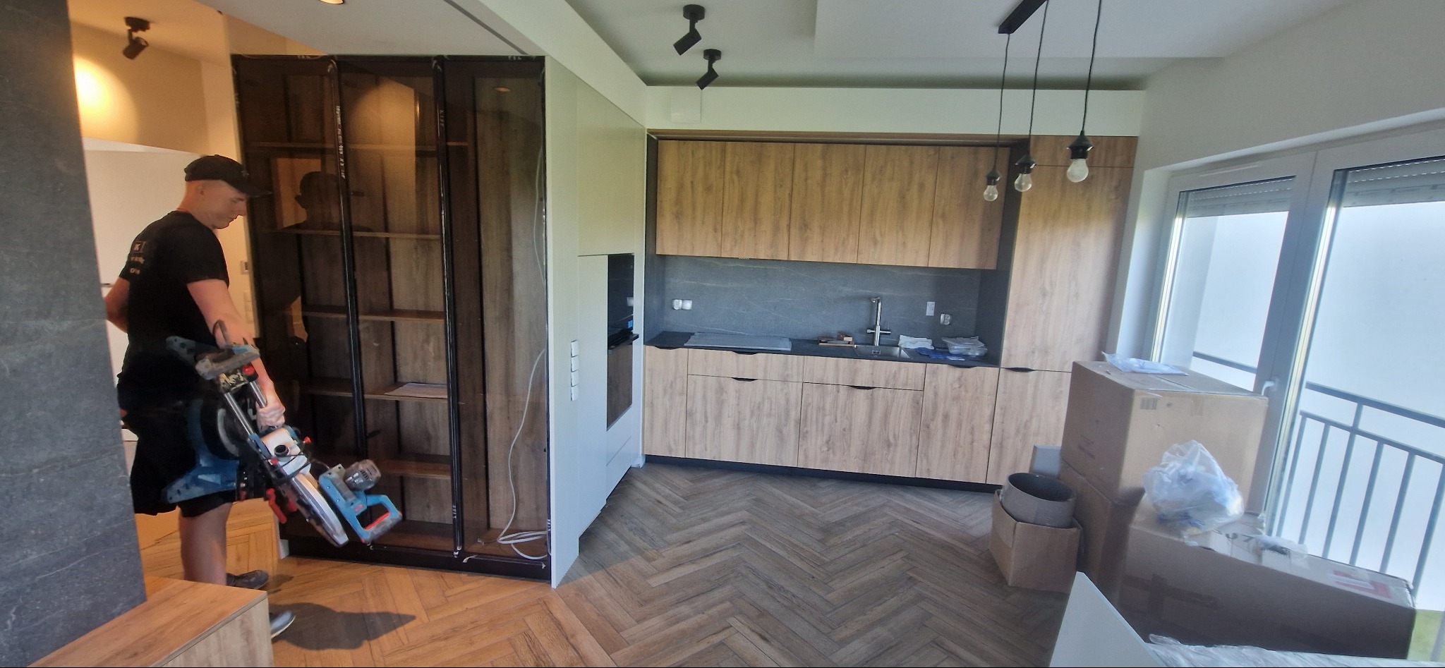 Montaż zabudowy meblowej: mężczyzna z piłą ukosową przy szafie z półkami na tle nowej kuchni z drewnianymi frontami i parkietem w jodełkę.