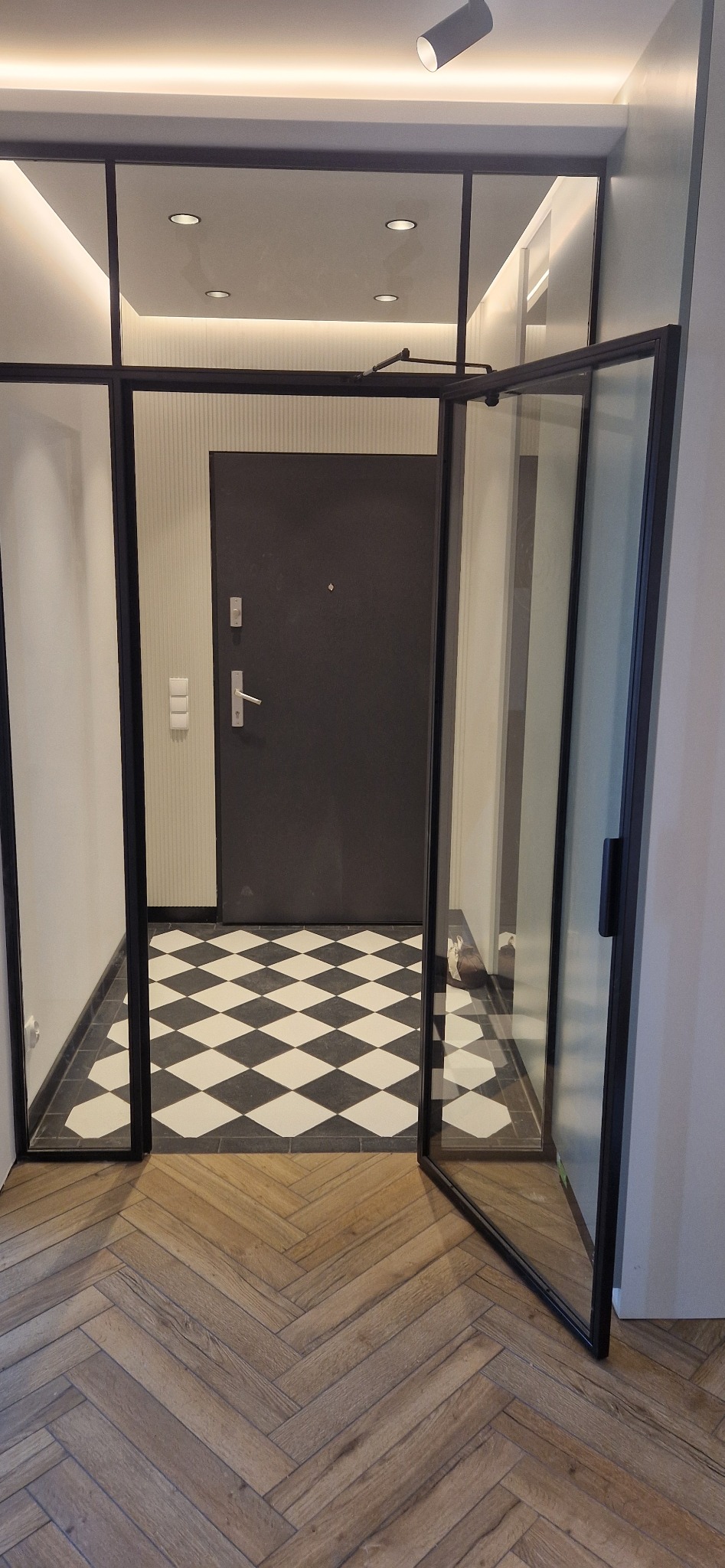 Nowoczesne wnętrze z otwartymi szklanymi drzwiami w czarnej ramie, ukazujące podłogę w jodełkę i czarno-białą szachownicę w przedpokoju.