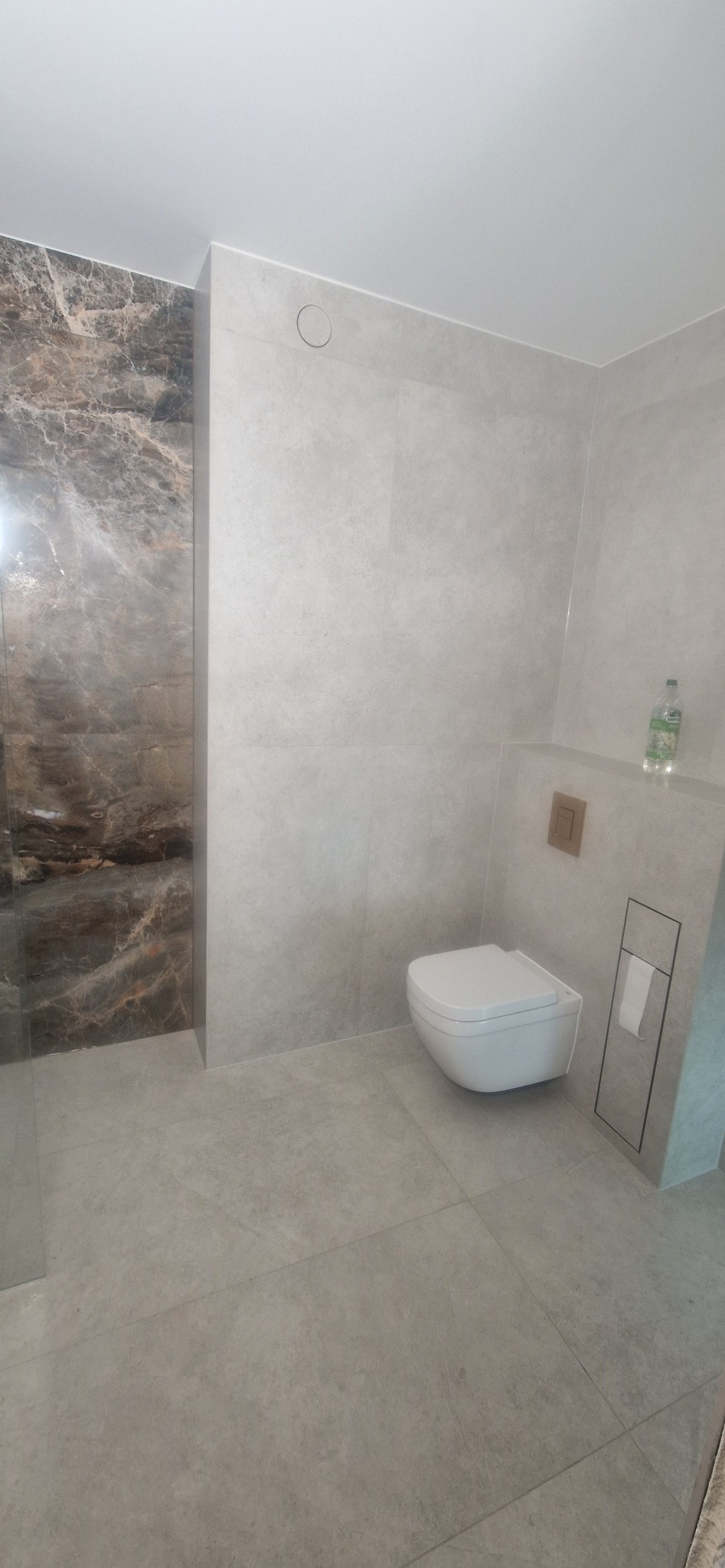 Nowoczesna łazienka po remoncie: szare płytki imitujące kamień, podwieszana toaleta, marmurowa ściana pod prysznicem, minimalistyczny design.