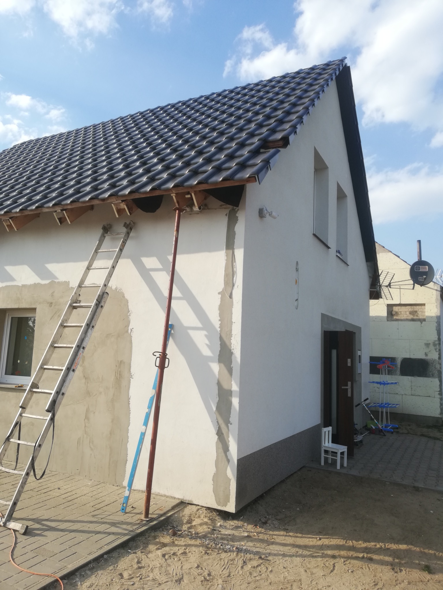 Dom murowany w trakcie wykończenia elewacji, widoczne rusztowanie, drabina oparta o ścianę, fragment położonej dachówki i ocieplenie budynku obok.