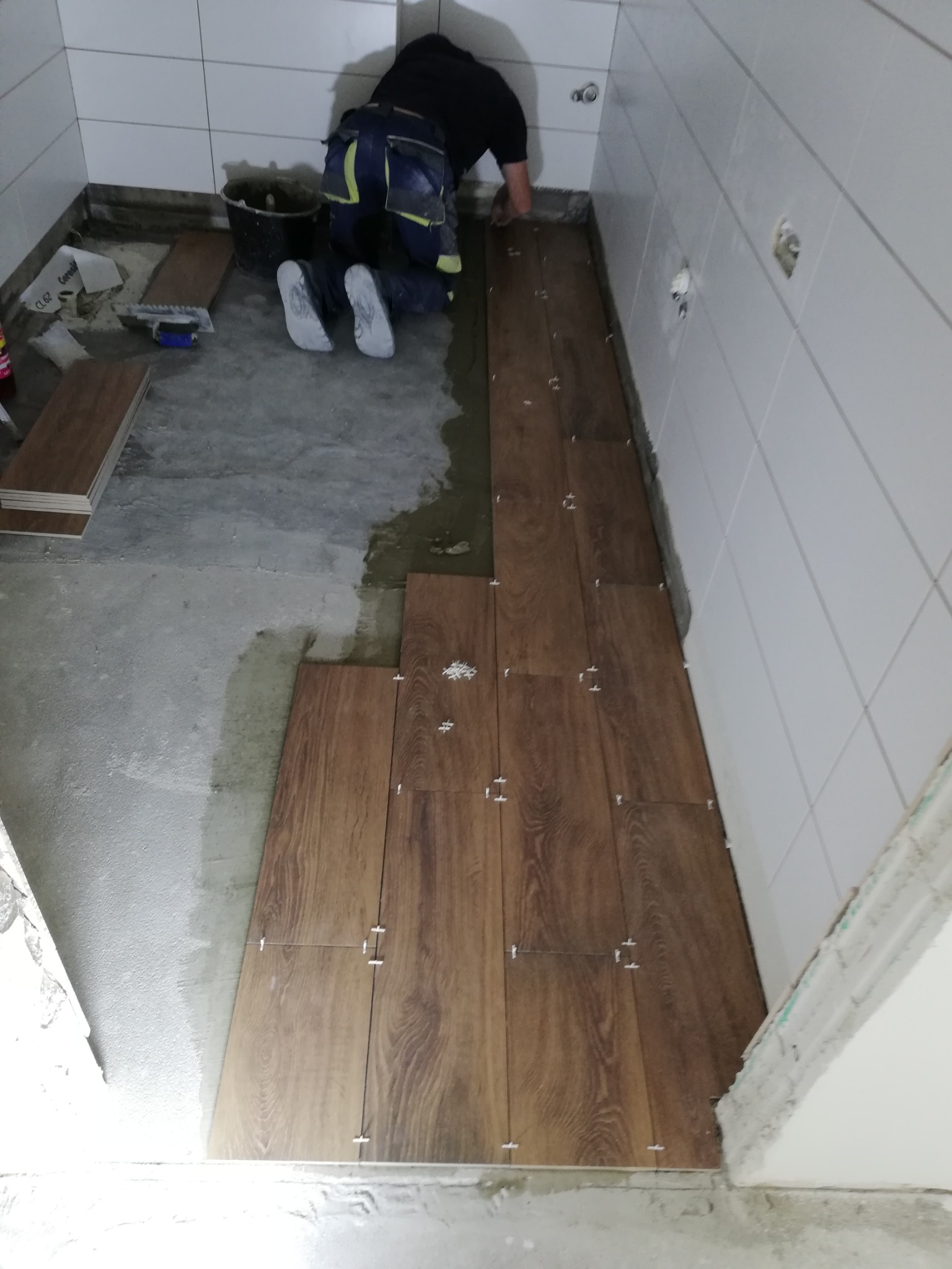 Układanie brązowych płytek podłogowych imitujących drewno w trakcie remontu łazienki, widoczne białe płytki ścienne, krzyżyki dystansowe oraz pracownik w odzieży roboczej.