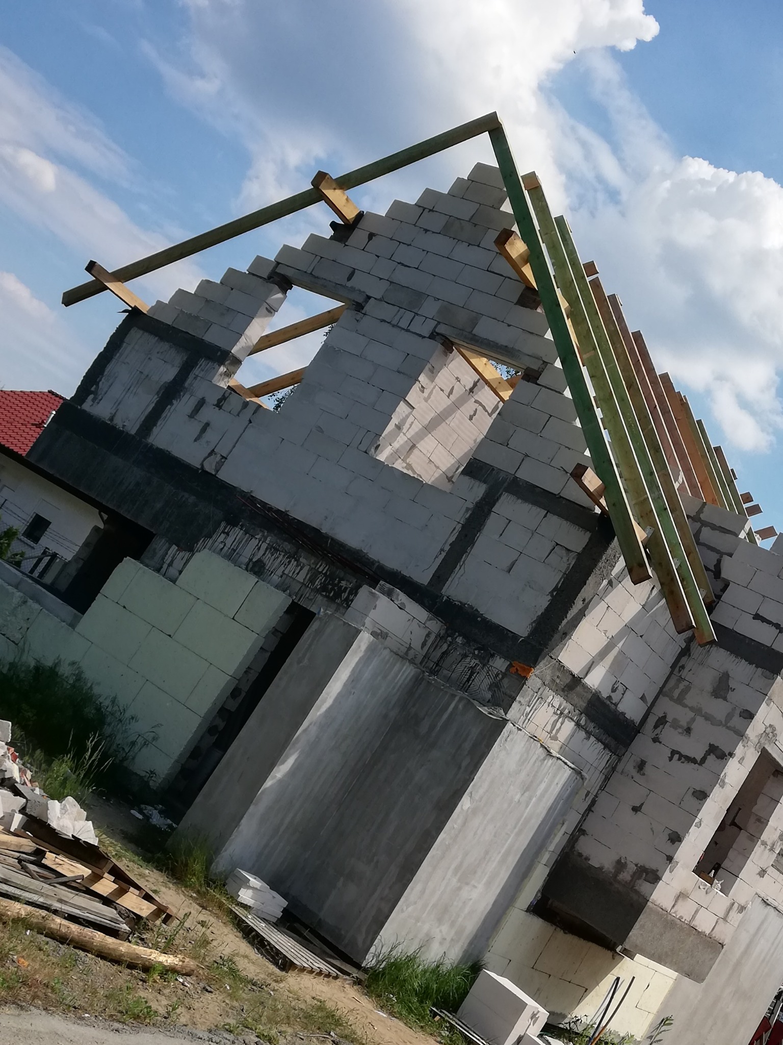 Wznoszenie ścian domu z bloczków betonowych, widoczne drewniane elementy konstrukcji dachu na tle błękitnego nieba z chmurami.
