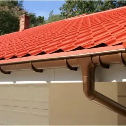 Czerwona dachówka ceramiczna na dachu domu z brązowymi rynnami, garaż w tle. Widok z dołu, oświetlenie dzienne.