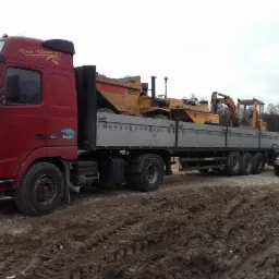 Czerwona ciężar&oacute;wka z platformą transportową przewozi ż&oacute;łty walec drogowy i koparkę na błotnistym terenie, obok stoi ciemny samoch&oacute;d osobowy.