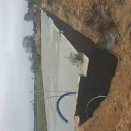 Wylany, mokry beton na płycie fundamentowej w wykopie, widoczne ściany wykopu z ziemi, niebieski przewód i metalowy drut na pierwszym planie, w tle drzewa i ogrodzenie.