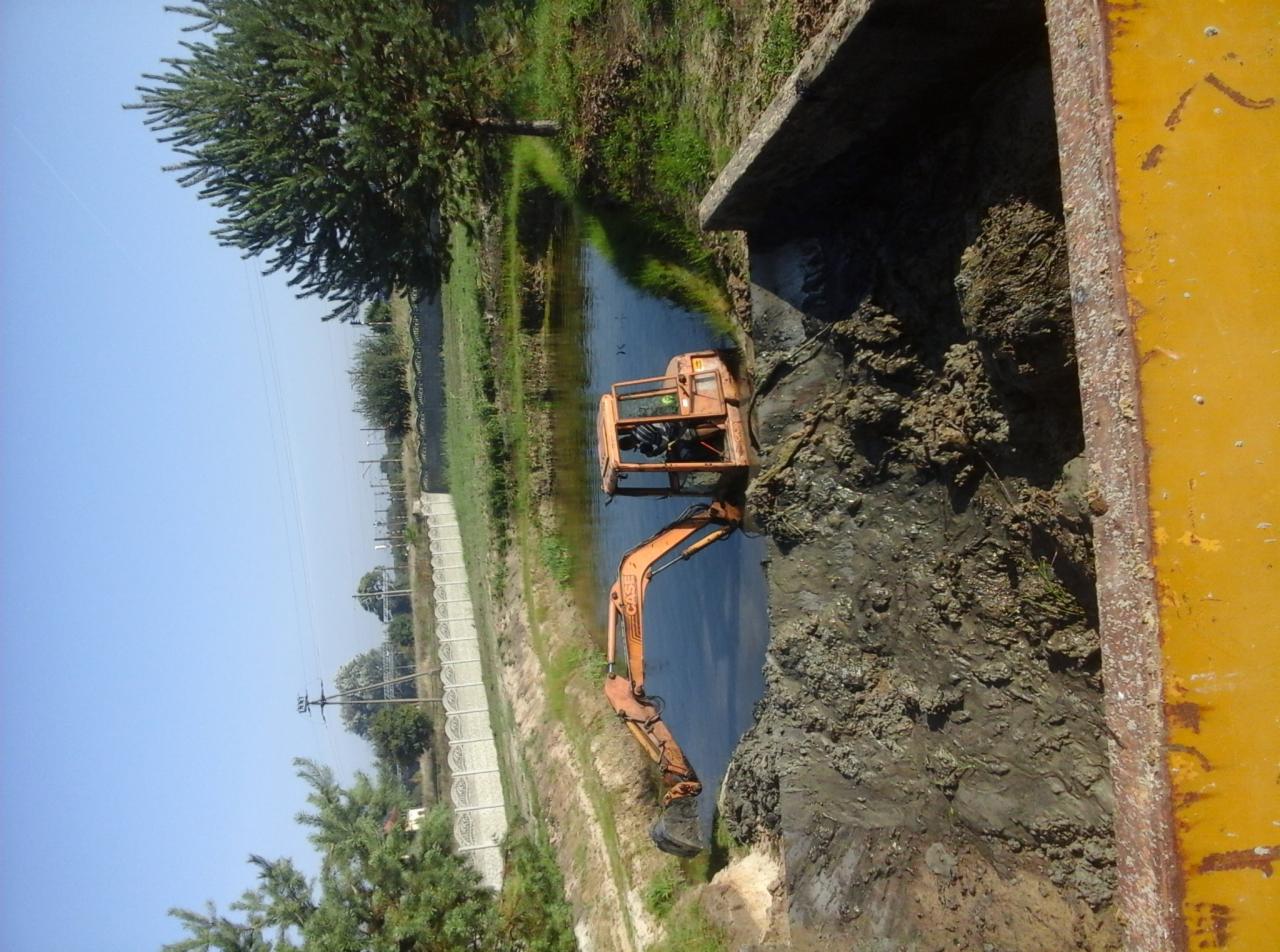 Pomarańczowa koparka CASE w kanale wodnym, czyszczenie rowu melioracyjnego. Widok z góry, z fragmentem żółtej burty ciężarówki. Prace ziemne w terenie podmokłym.