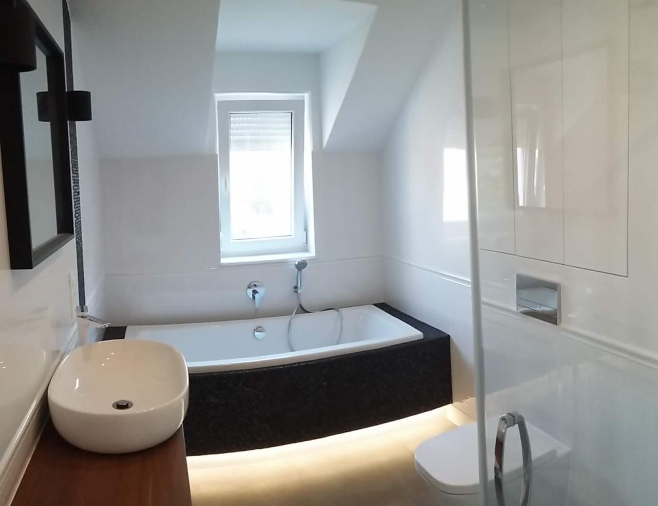 Nowoczesna łazienka z wolnostojącą wanną obudowaną czarnym kamieniem, owalną umywalką na drewnianym blacie i podświetleniem LED pod wanną, widok na okno i toaletę za szklaną ścianką.