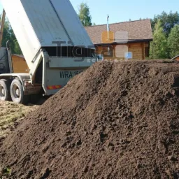 Ciężarówka z otwartą skrzynią wyładowuje ciemną ziemię na trawnik przed domami, widoczny numer rejestracyjny WRA 164.
