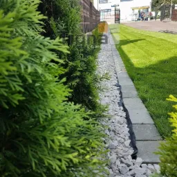 Ścieżka w ogrodzie wyłożona szarym żwirem, oddzielona obrzeżem z szarych kostek, obok rosną tuje i trawa, w tle budynek.