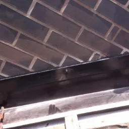 Detal dachu: czarna obróbka blacharska przy kominie z ciemnej cegły klinkierowej, widok z dołu. Elementy konstrukcji drewnianej w tle.