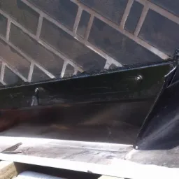 Detal wykończenia komina z cegły klinkierowej na dachu, z czarną obróbką blacharską. Widoczne fragmenty dachówek i papy.