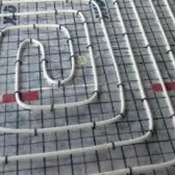 Białe rury ogrzewania podłogowego ułożone spiralnie na czarnej siatce z czerwonymi znacznikami, mocowane czarnymi klipsami, widok z góry, oświetlenie naturalne.