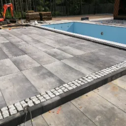 Nowoczesna przestrzeń wokół basenu z ułożoną szarą kostką brukową i obrzeżem z białej kostki granitowej; w tle minikoparka i zwinięta trawa, częściowo wypełniony basen z niebieską wodą.