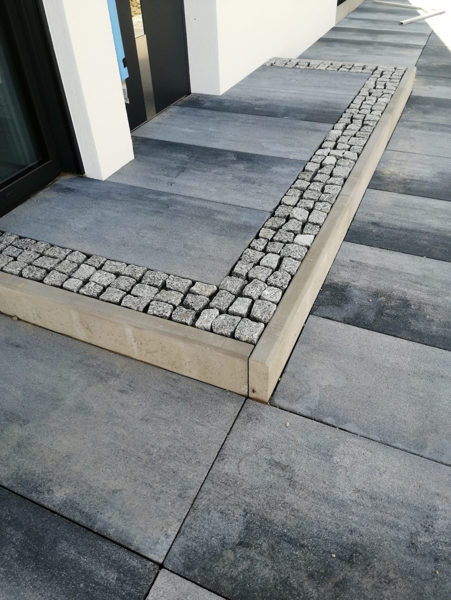 Szare, prostokątne płyty chodnikowe z bordiurą z kostki granitowej i betonowym obrzeżem przy wejściu do budynku z ciemnymi oknami.