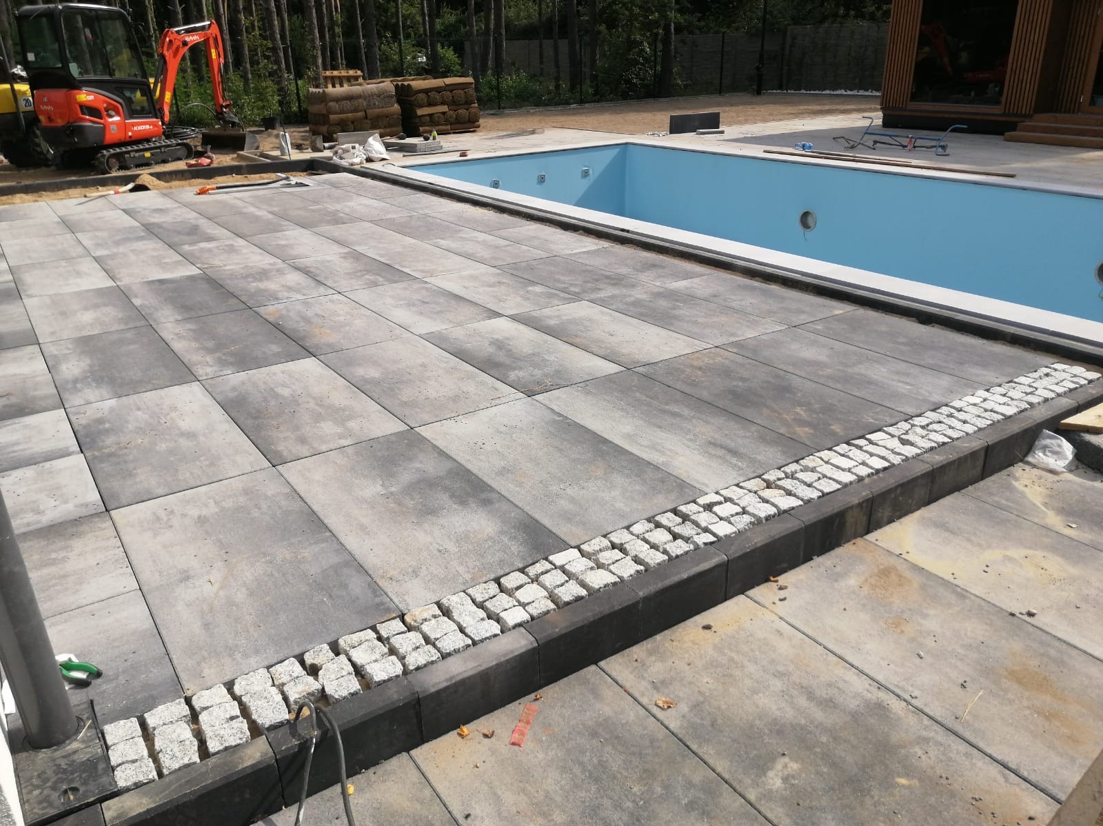Nowoczesna przestrzeń wokół basenu z ułożoną szarą kostką brukową i obrzeżem z białej kostki granitowej; w tle minikoparka i zwinięta trawa, częściowo wypełniony basen z niebieską wodą.