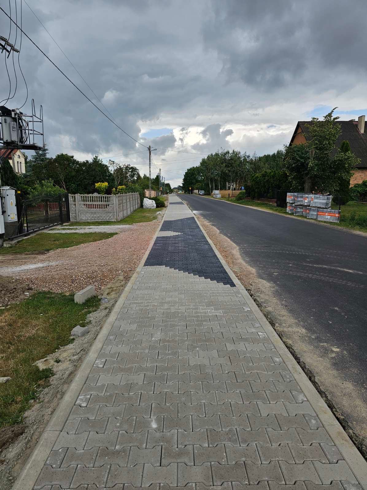 Ułożona chodnikowa kostka w kształcie litery I, z fragmentem czarnej kostki w trakcie układania, widok na ulicę w pochmurny dzień.