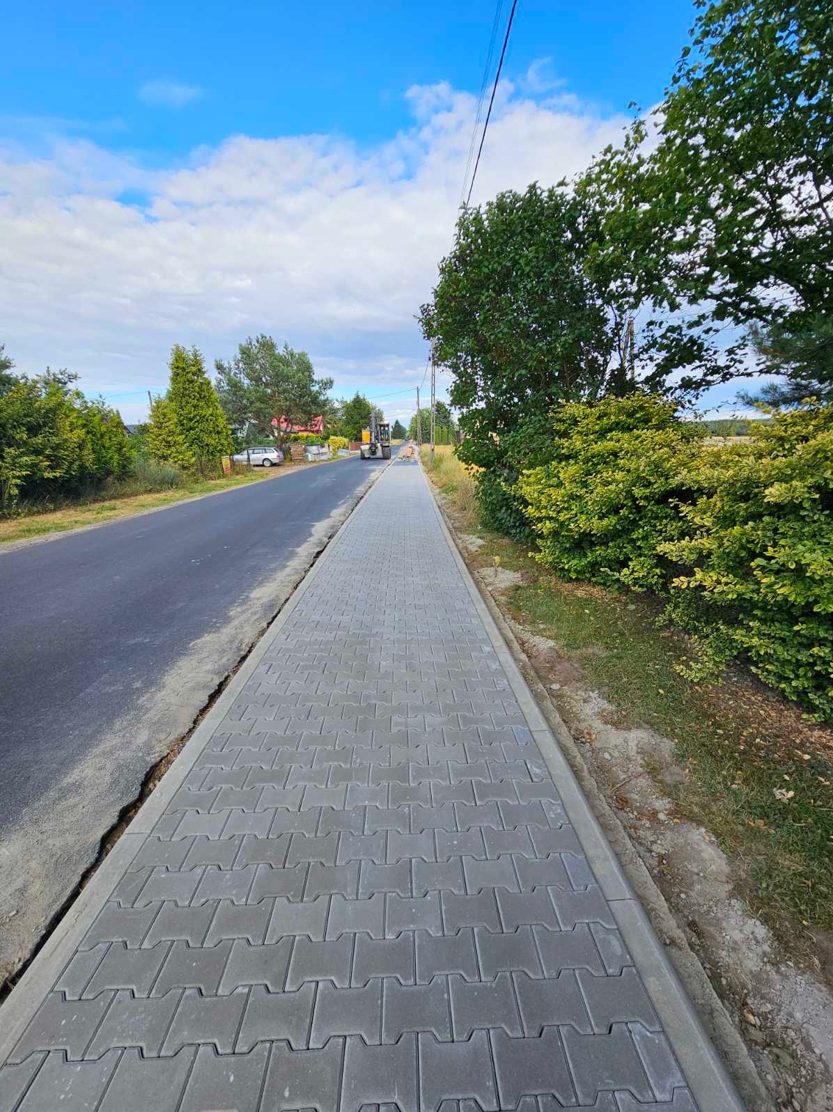 Ułożona szara kostka brukowa tworząca chodnik wzdłuż drogi asfaltowej, z widoczną maszyną budowlaną w oddali i zielenią po bokach, sfotografowana w słoneczny dzień.