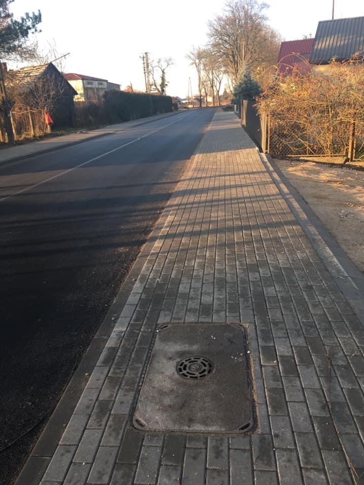 Ułożona szara kostka brukowa tworząca chodnik wzdłuż asfaltowej drogi, z widoczną studzienką kanalizacyjną i domami w tle, oświetlona popołudniowym słońcem rzucającym długie cienie.