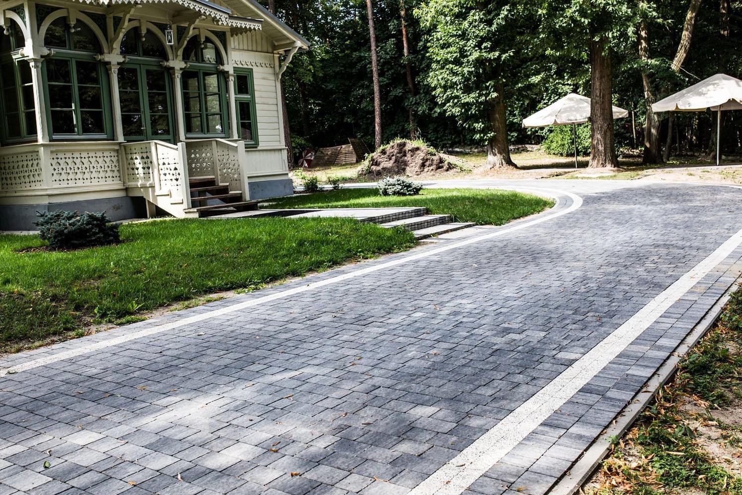 Ułożona szara kostka brukowa na podjeździe przed domem w stylu retro z zielonymi oknami, białe obrzeża, trawnik i parasole ogrodowe w tle.