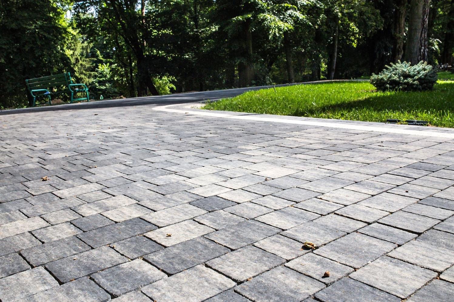 Ułożona kostka brukowa w odcieniach szarości tworząca ścieżkę w ogrodzie, widoczna zielona trawa, krzew i ławka w tle.