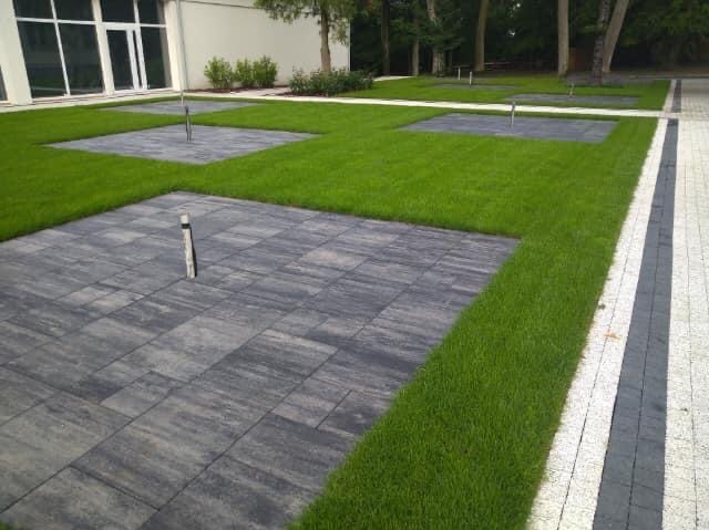 Nowoczesna aranżacja terenu z szarej kostki brukowej ułożonej w kwadraty otoczone trawnikiem, z widocznymi słupkami oświetleniowymi, w tle budynek z dużymi oknami.