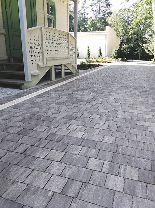 Ułożona, szara kostka brukowa prowadząca do domu z białym gankiem i zielonymi drzwiami, oddzielona białym pasem od trawnika z iglakami.