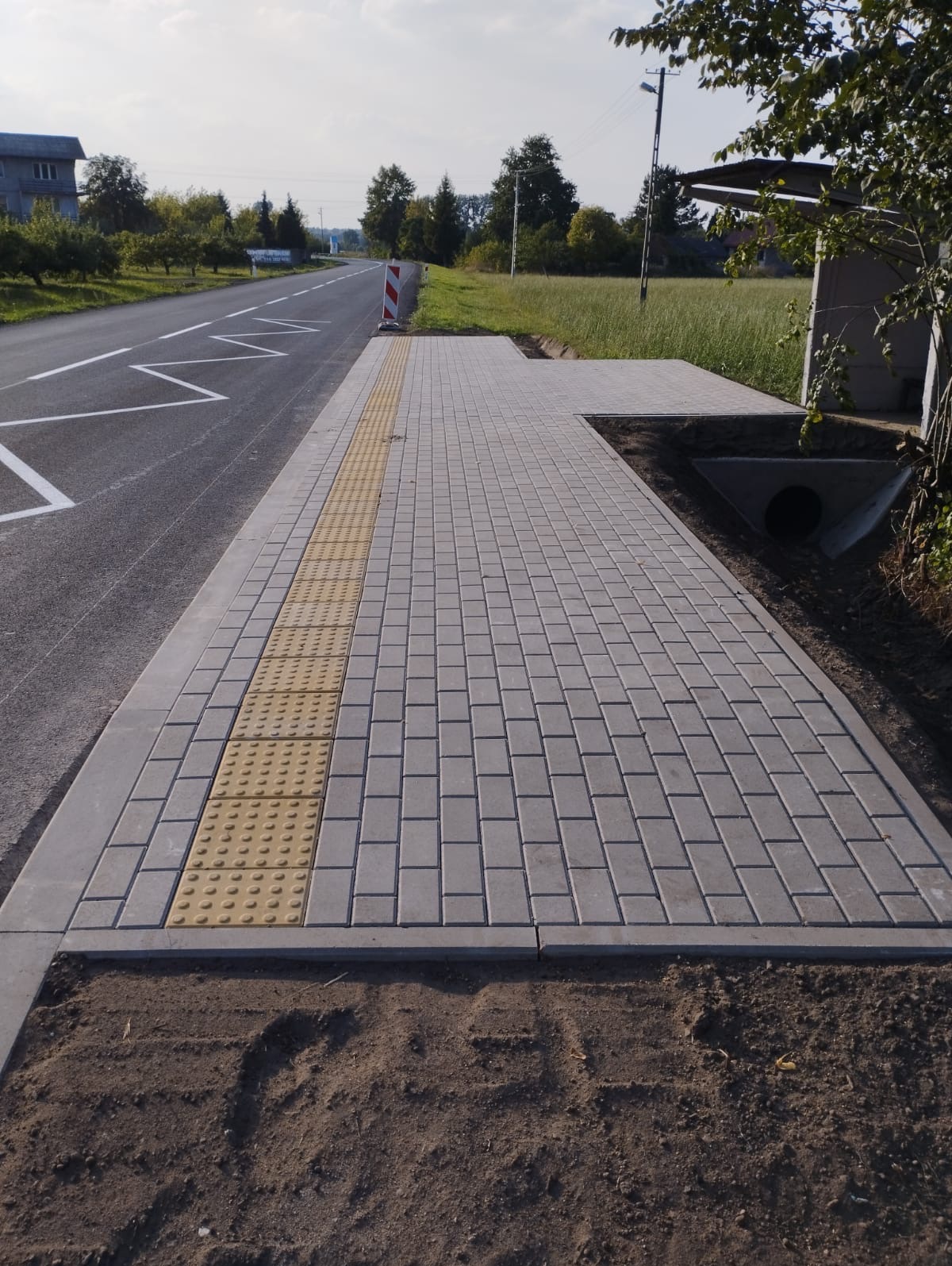 Nowo ułożony chodnik z szarej kostki brukowej z żółtymi płytami sensorycznymi dla osób niewidomych, przylegający do jezdni z widocznym oznakowaniem poziomym, w tle budynki i drzewa.