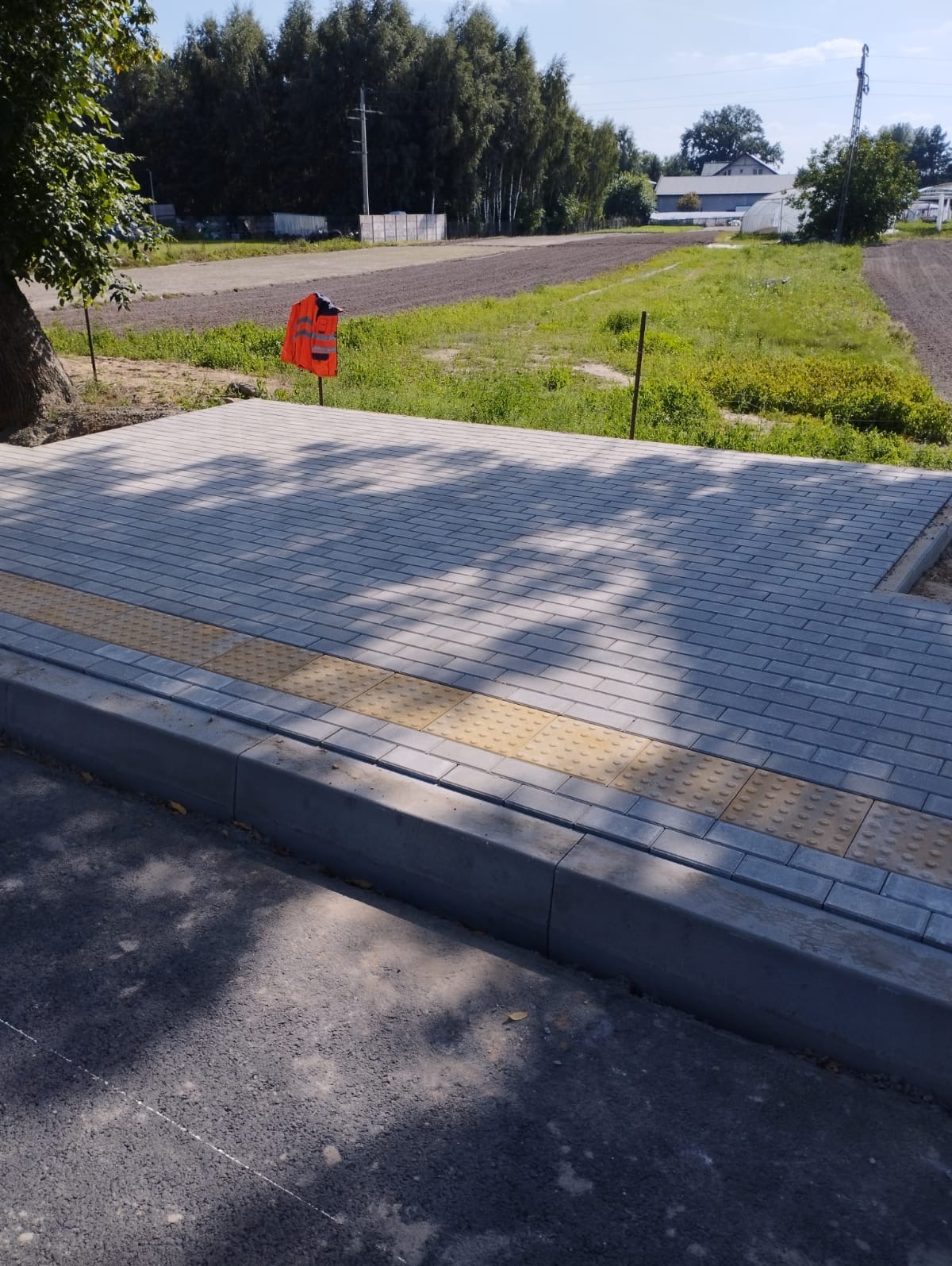 Nowo ułożona, szara kostka brukowa z żółtymi płytami dotykowymi dla osób niedowidzących, obrzeża betonowe, w tle pole i drzewa, pomarańczowa kamizelka odblaskowa na tyczce.