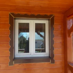 Drewniana elewacja domu z oknem w białej ramie z dekoracyjnym, brązowym obramowaniem.