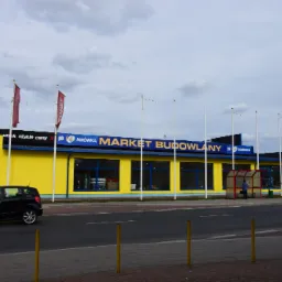 Żółto-niebieski budynek marketu budowlanego Mrówka z reklamami Neonet, widziany z ulicy w Hajnówce, z przejeżdżającym czarnym samochodem.