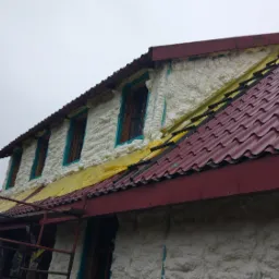 Poddasze domu jednorodzinnego w trakcie ocieplania pianą PUR, widoczne okna z turkusową stolarką okienną, fragment czerwonej blachodachówki i rusztowania.
