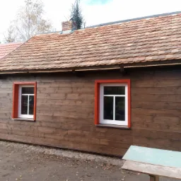 Drewniany dom z nową dachówką ceramiczną typu karpiówka, pomalowanymi na czerwono obramowaniami okien i brązową elewacją z desek.