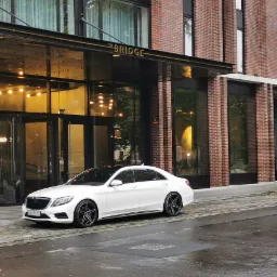 Biały Mercedes S-klasy zaparkowany przed nowoczesnym budynkiem z cegły z napisem 'The Bridge' nad wejściem, mokra nawierzchnia ulicy.