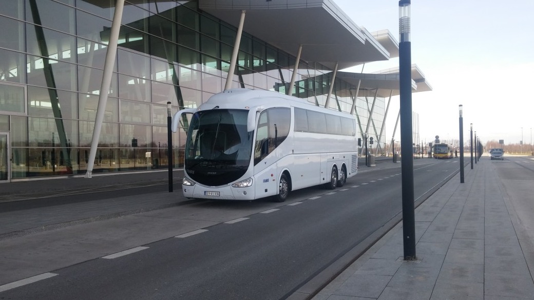 Biały autokar turystyczny marki Irizar zaparkowany przed nowoczesnym budynkiem z przeszkloną fasadą i zadaszeniem wspartym na białych kolumnach. Na pierwszym planie asfaltowa droga z białymi liniami.