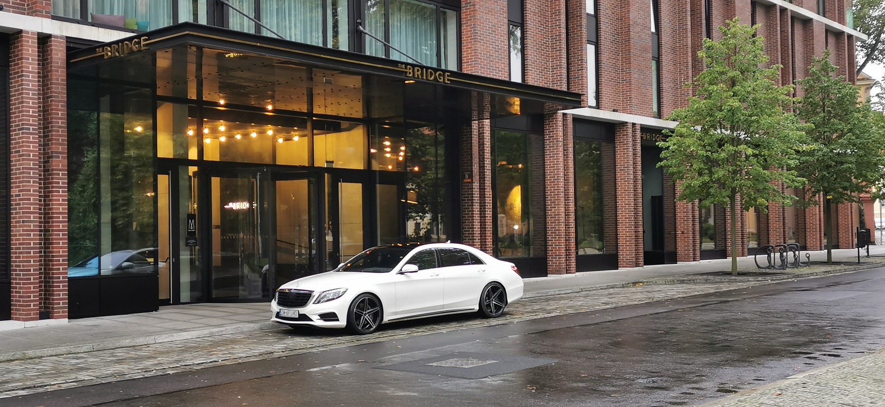 Biały Mercedes S-klasy zaparkowany przed nowoczesnym budynkiem z cegły z napisem 'The Bridge' nad wejściem, mokra nawierzchnia ulicy.