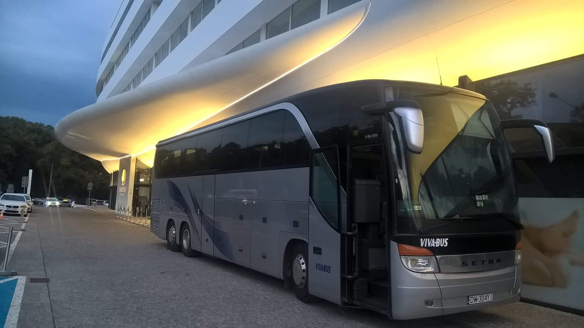 Nowoczesny autokar VIVA-BUS Setra zaparkowany przed futurystycznym budynkiem hotelu z oświetlonym baldachimem. Otwarte drzwi autobusu.
