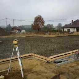 Przygotowanie zbrojenia i szalunku pod płytę fundamentową na gruncie, widoczny niwelator laserowy na statywie, w tle domy jednorodzinne i hałda ziemi.