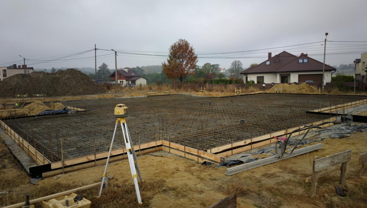 Przygotowanie zbrojenia i szalunku pod płytę fundamentową na gruncie, widoczny niwelator laserowy na statywie, w tle domy jednorodzinne i hałda ziemi.