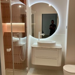 Wykończenia Wnętrz Łukasz Pacia - Nowoczesna łazienka z okrągłym lustrem podświetlanym LED, białą umywalką nablatową na szafce z frezowanymi frontami i kabiną prysznicową z brązowymi płytkami w tle. W lustrze odbija się osoba...