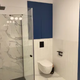 Nowoczesna łazienka z kabiną prysznicową wykończoną płytkami imitującymi marmur, wiszącą toaletą i szafką w kolorze granatowym, akcenty w kolorze czarnym.